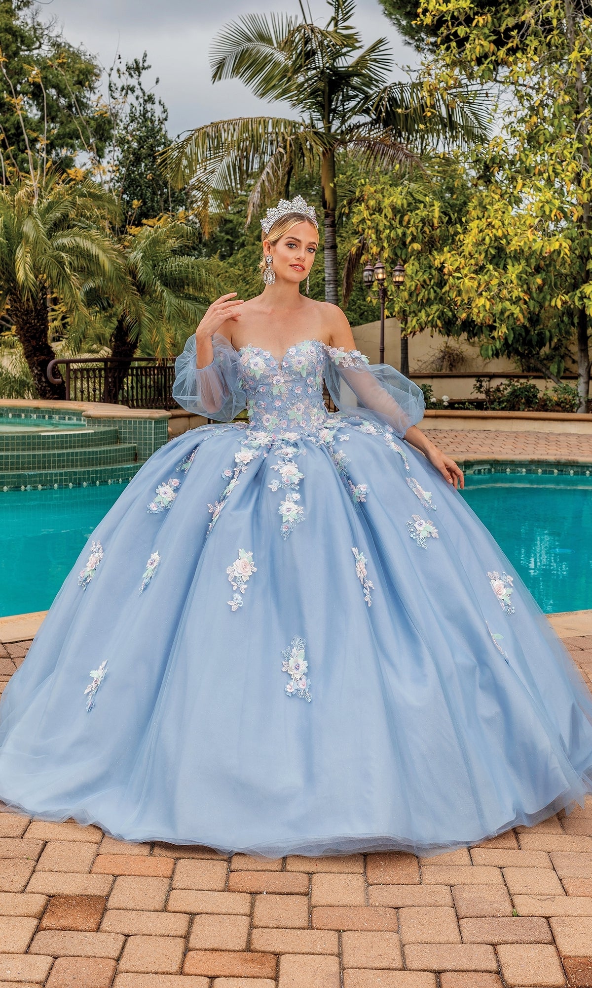 A woman poses by a pool surrounded by greenery, wearing the Quinceanera Dress 1871 By Dancing Queen—an A-line ball gown with lace floral applique and puffy sleeves.
